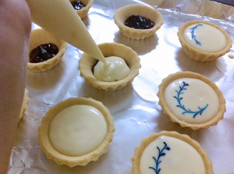 Close-up of small pastry tart shells being filled with cream and jam, some decorated with blue icing designs, resting on a foil surface; soft pastel pinks, creams, and pale blues in a warm, home-kitchen mood.